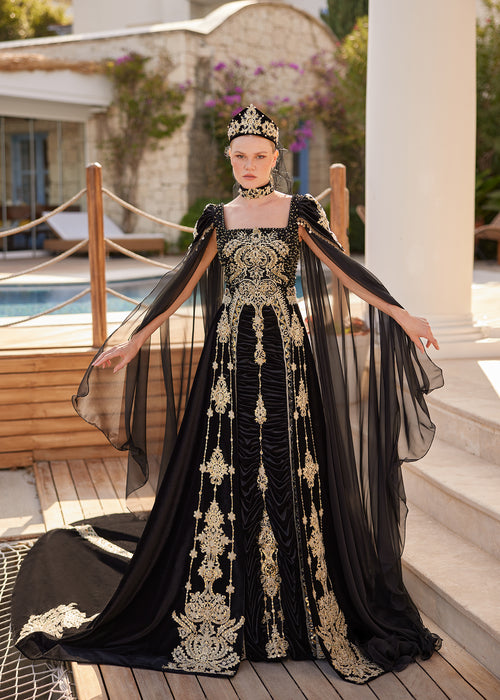 Front view of the black cape sleeve square neck formal kaftan gown with gold crystal embroideries, matching crown headpiece, and choker necklace by Sultan Dress.