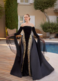 Front view of the black draped boat-neck mermaid formal gown styled with the open detachable overskirt and gold sequin embroidery.