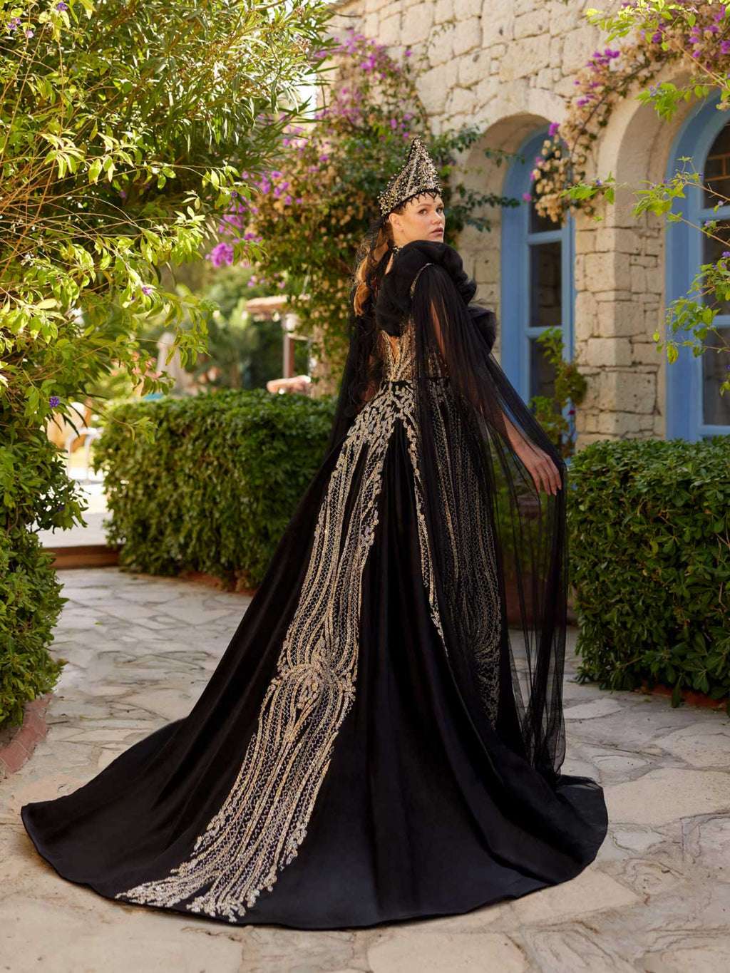 Rear view showing the dramatic flowing tulle shoulder cape and fitted back pattern of the black velvet gold sequin henna dress.