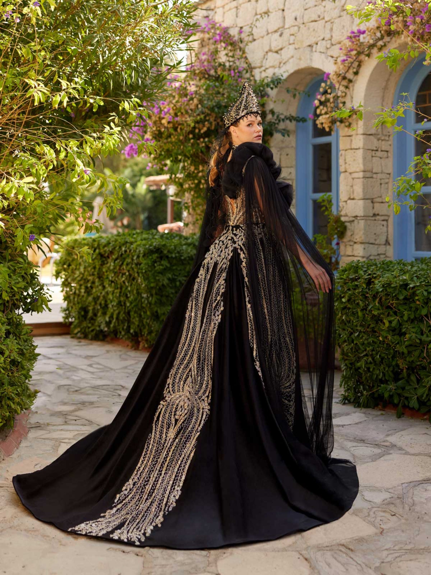 Rear view showing the dramatic flowing tulle shoulder cape and fitted back pattern of the black velvet gold sequin henna dress.