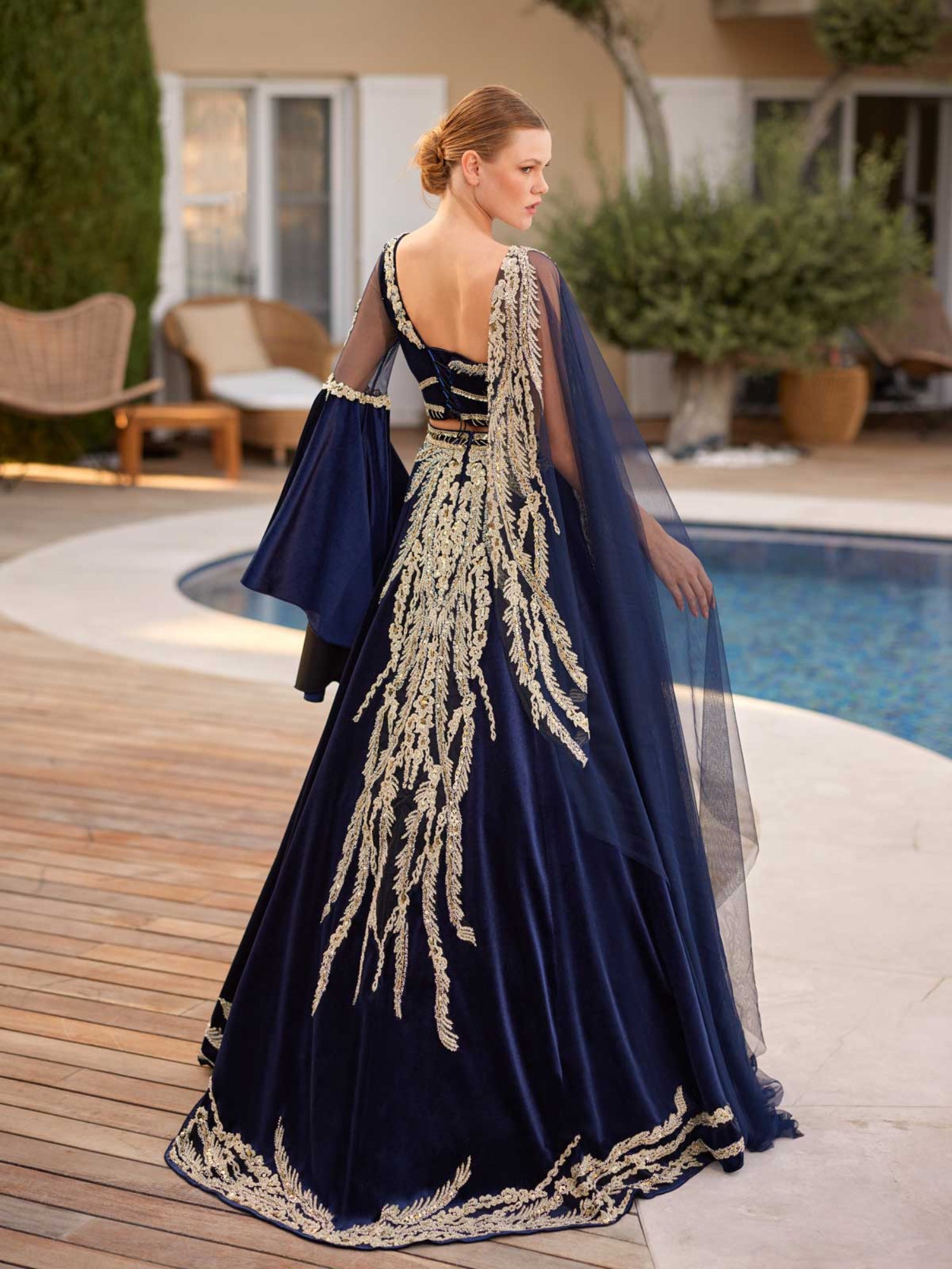 Rear view showing the scoop back bodice, gold embroidered skirt train, and draped sheer navy cape on the velvet caftan gown.