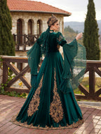Rear view showing the gold-embroidered skirt panels, crown accessory, and layered lace bell sleeves on the emerald green velvet kaftan gown.
