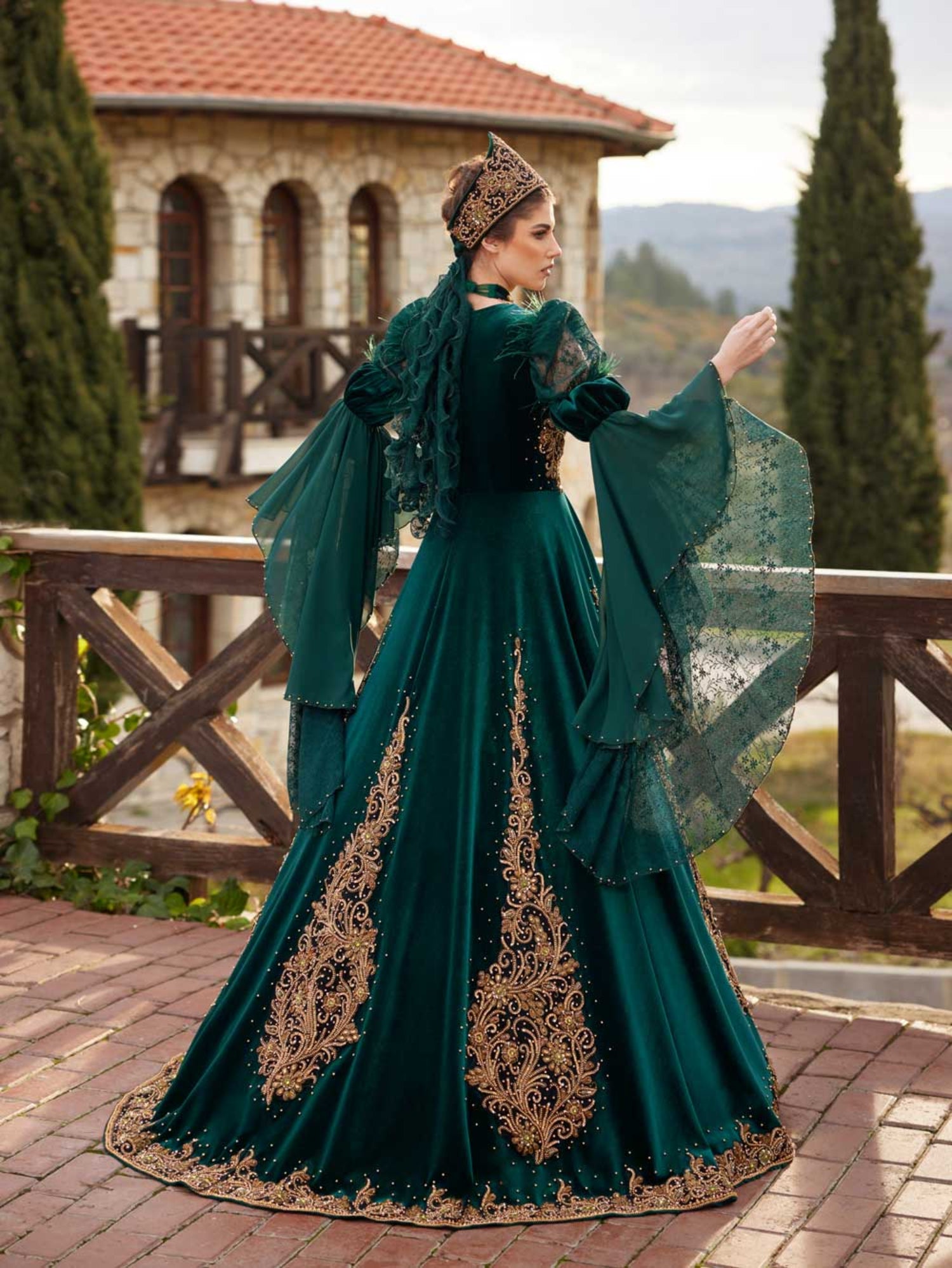 Rear view showing the gold-embroidered skirt panels, crown accessory, and layered lace bell sleeves on the emerald green velvet kaftan gown.