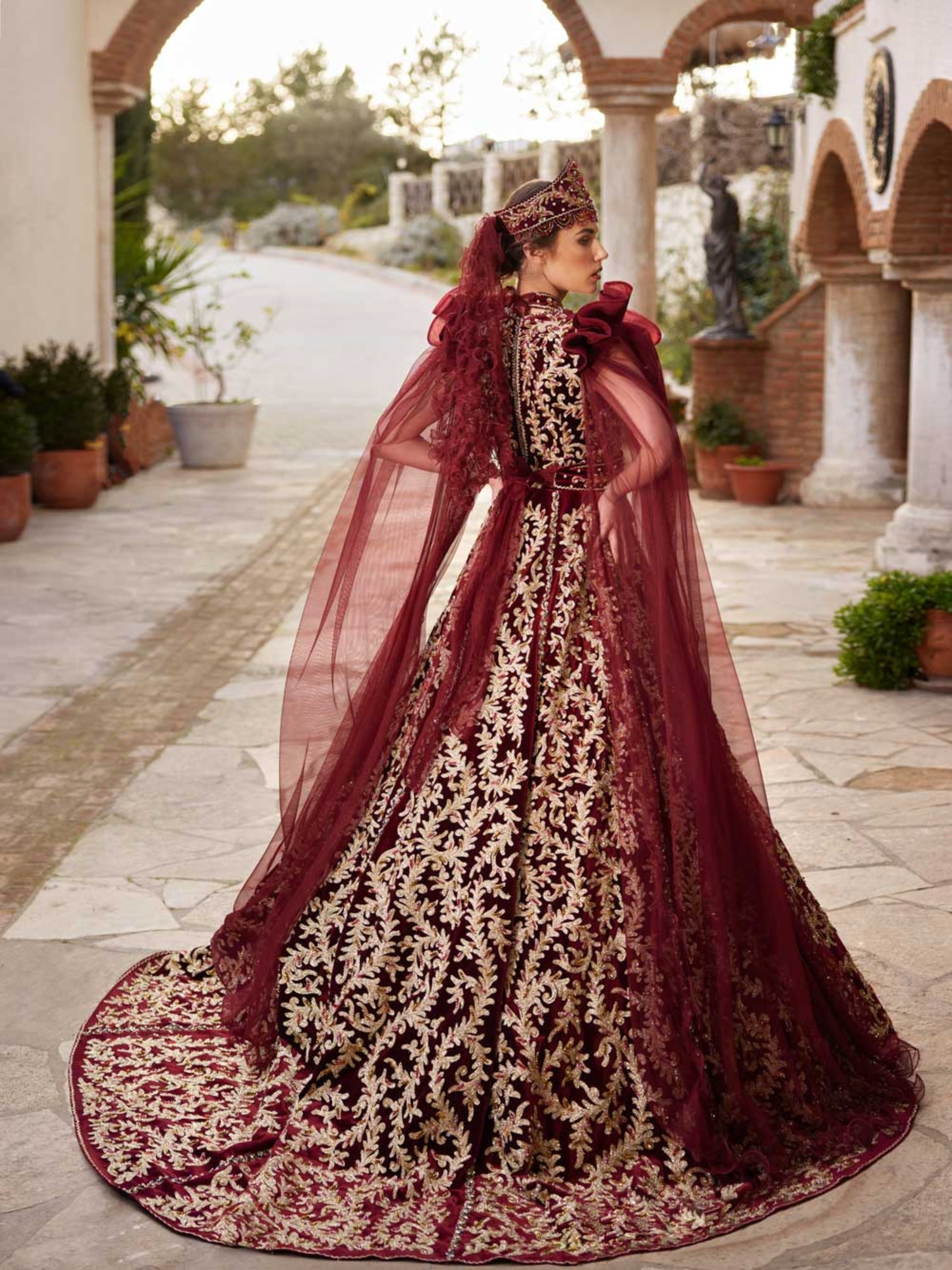 Rear view showing the gold-embroidered train, crown headpiece, and ruffled mesh shoulder cape on the maroon velvet bridal gown.